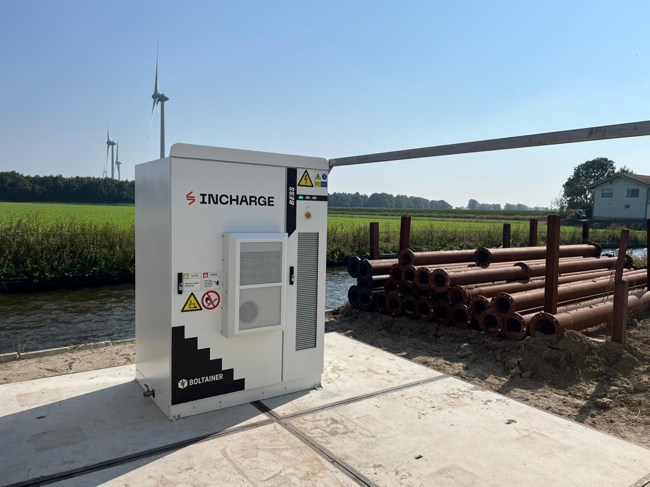 Outdoor battery storage cabinet beside a polder waterway with wind turbines
